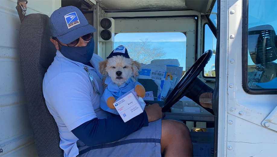 mail man sitting in his truck while holding a white dog who has its paws on a white delivery box
